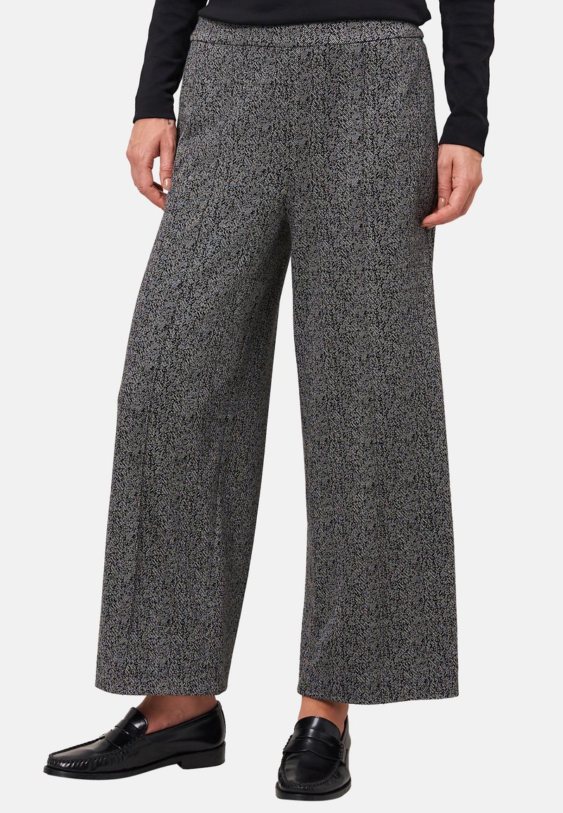 Persona che indossa pantaloni larghi grigi a motivo houndstooth con mocassini in pelle nera e una maglia nera a maniche lunghe, in piedi davanti a uno sfondo neutro.