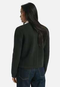 Kvinna med långt mörkt hår som bär en svart texturerad jacka och blå denimjeans, vänd mot vänster med rygg- och sidoprofil synlig.