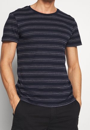 Hombre con camisa azul marino de manga corta con rayas horizontales blancas y pantalones negros, manos en los bolsillos, de pie frente a un fondo claro.