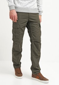 Olivgrüne Cargo-Shorts mit mehreren Taschen, aus strapazierfähigem Stoff; kombiniert mit braunen Schnürsneakern. Die Hose hat eine lockere Passform.