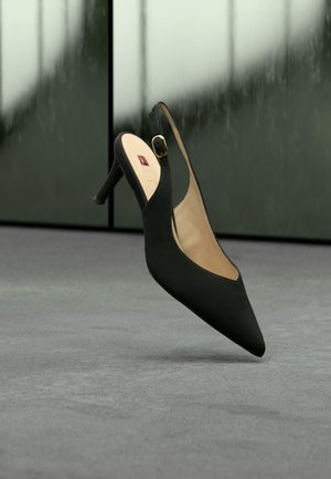 Zapato de tacón alto tipo slingback negro con punta afilada y pequeña hebilla dorada, mostrado flotando sobre una superficie gris con un fondo de vidrio desenfocado.