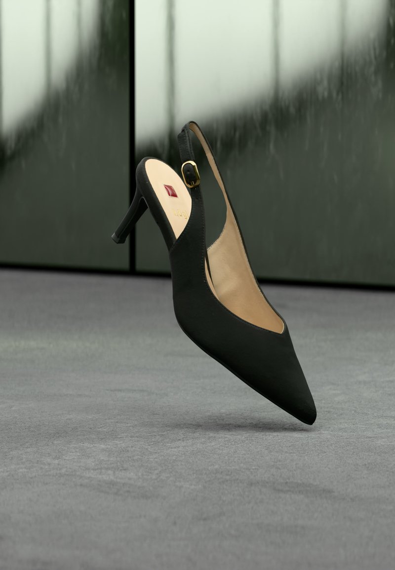 Μαύρο γυναικείο παπούτσι με λουράκι πίσω και ψηλό τακούνι, μυτερή μύτη και μικρή χρυσή αγκράφα, που αιωρείται πάνω από γκρι επιφάνεια με θολό γυάλινο φόντο.