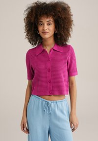 Fuksijas krāsas adīts īsām piedurknēm kardigans ar podziņu aizdari un klasisku apkakli, apvienots ar gaiši zilu biksēm ar aukliņu.