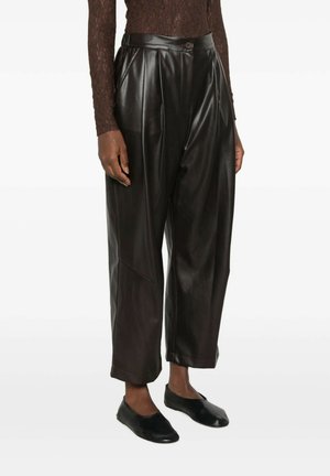 Donna che indossa pantaloni neri in ecopelle a vita alta con pieghe frontali, abbinati a scarpe basse nere e una maglia marrone a maniche lunghe.