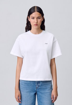 T-shirt bianca a maniche corte in cotone con scollo rotondo e un piccolo logo nero sul petto, abbinata a jeans in denim azzurri.