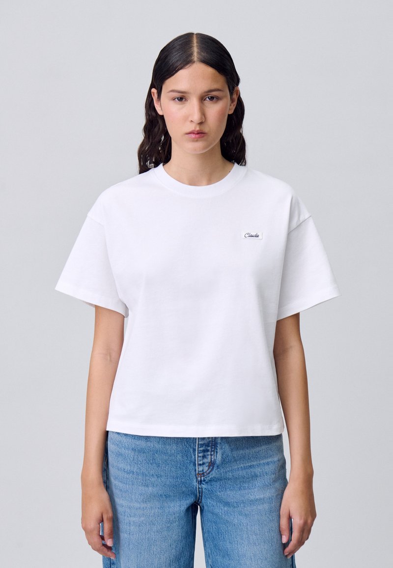 T-shirt bianca a maniche corte in cotone con scollo rotondo e un piccolo logo nero sul petto, abbinata a jeans in denim azzurri.