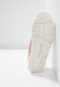 Sneaker Reebok con suola esterna cremosa texturizzata, tomaia beige e dettagli rosa. Il design comprende marchio visibile e materiali stratificati.