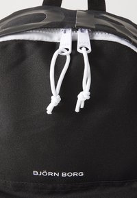 Mochila de tecido preta com zíperes e cordas brancas. Apresenta um logotipo e o nome da marca "BJÖRN BORG" impressos em branco na parte frontal.