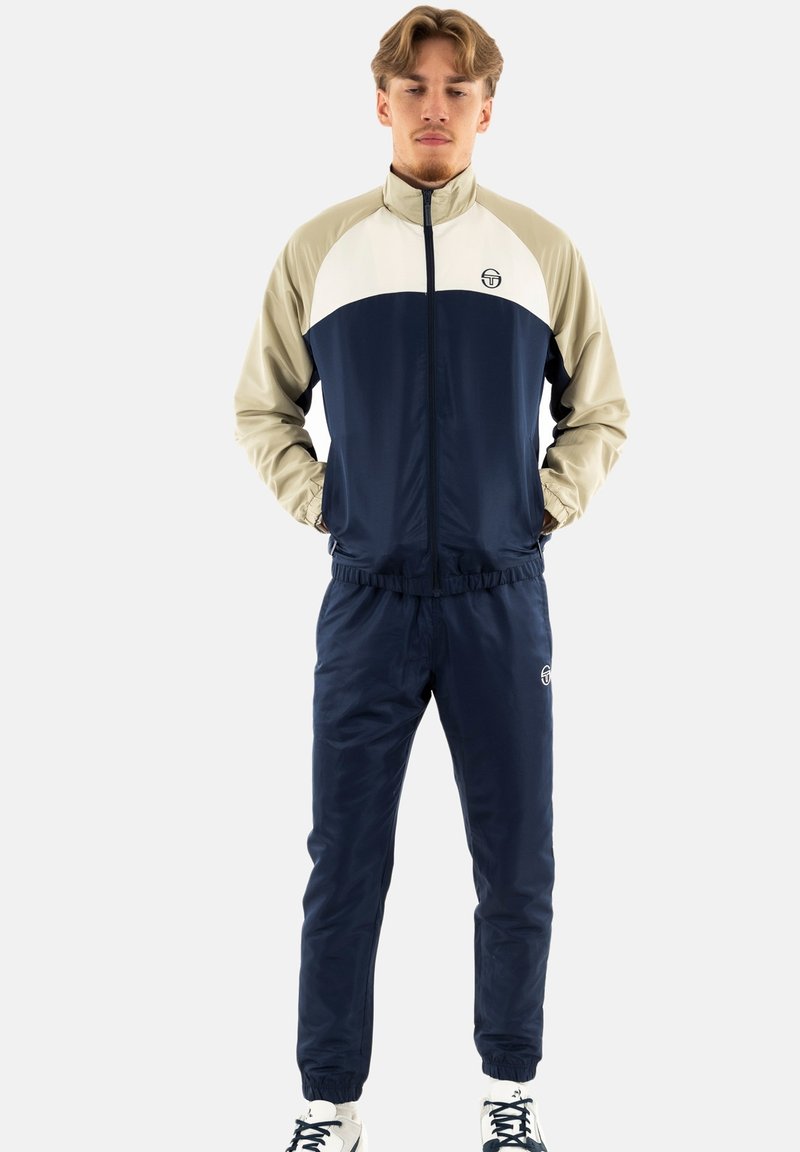 Marineblauer und beiger Trainingsanzug bestehend aus einer mit Reißverschluss versehenen Jacke mit hohem Kragen und elastischen Bundhosen. Hergestellt aus glattem, leichtem Material.