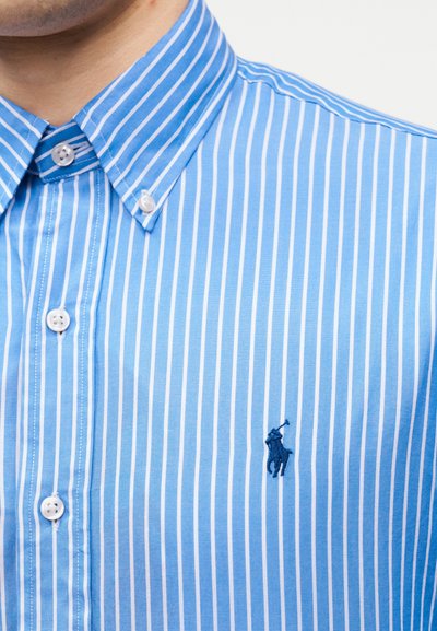 Πουκάμισο με κάθετες μπλε και άσπρες ρίγες, με κουμπιά και γιακά με κουμπιά, και σκούρο μπλε λογότυπο παίκτη πόλο στο στήθος.