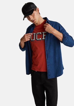 Blaues Jeanshemd über einem roten Grafik-T-Shirt mit UCE-Schriftzug, kombiniert mit schwarzer Hose und schwarzer Lederkappe.