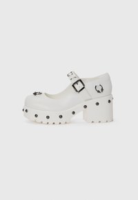 White platform shoes with a rounded toe, buckle strap, and circular studs. Textured sole with a slight heel and decorative hardware detail.