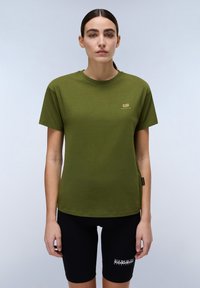 Olivengrünes Baumwoll-T-Shirt mit kurzen Ärmeln, Rundhalsausschnitt und kleinem goldenen Logo auf der Brust. Kombiniert mit schwarzen Fahrradhosen.