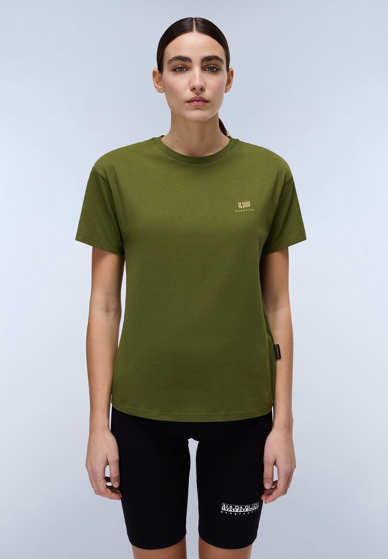 Olivengrünes Baumwoll-T-Shirt mit kurzen Ärmeln, Rundhalsausschnitt und kleinem goldenen Logo auf der Brust. Kombiniert mit schwarzen Fahrradhosen.