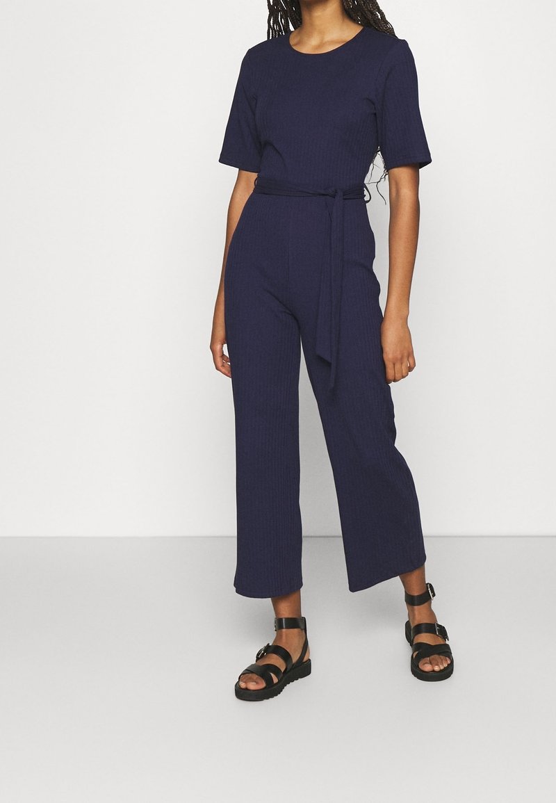 Marineblauer gestreifter Jumpsuit mit kurzen Ärmeln, Rundhalsausschnitt und tailliertem Design mit Gürtel. Kombiniert mit schwarzen Riemchensandalen. Flacher, weit geschnittener Stil.