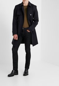 Schwarzer Trenchcoat mit Doppelreiheknöpfen, olivgrüner Unterhemd, dunkle Slim-Fit-Jeans und schwarze Lederschuhe. Glatte Textur, maßgeschneiderte Passform.