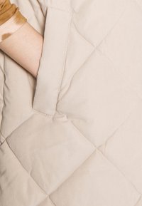 Gilet imbottito beige con un motivo cucito diagonale e tasca laterale, che mette in mostra un tessuto morbido e una consistenza liscia.