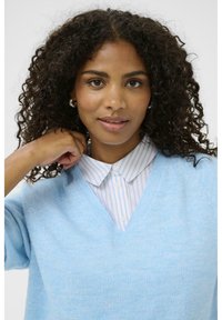 Gaiši zils v-veida džemperis, kas izgatavots no mīksta auduma, uzvilkts virs baltas krekla ar ziliem svītrām, ar brīvu piegriezumu.