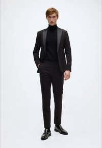 Zwart pak met satijnen revers, gedragen over een zwarte coltrui. Slim-fit broek en gepolijste zwarte leren schoenen. Effen achtergrond.