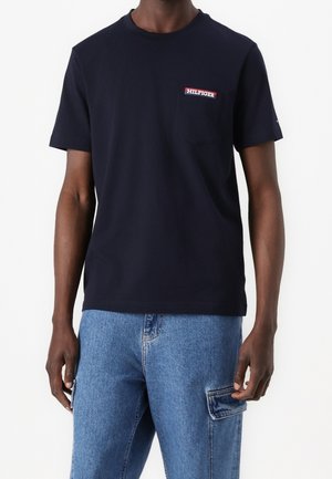 Mand iført en sort t-shirt med korte ærmer med et lille "Hilfiger" logo på venstre brystlomme og blå denim cargobukser.
