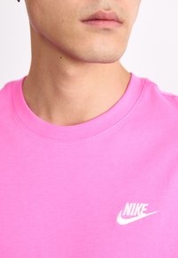 Rožinė trumpomis rankovėmis medvilninė marškinėliai su apvaliu kaklu ir siūlėmis, ant kairės pusės esantis baltas Nike logotipas.