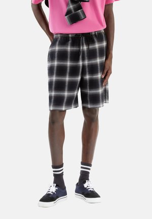 Schwarz-weiße Karotten-Shorts mit elastischem Bund und Kordelzug, aus leichtem Stoff gefertigt. Kombiniert mit marineblauen Turnschuhen und gestreiften Socken.