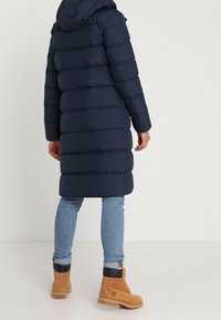 Marinblå vadderad jacka med horisontell sömnad, huva och knälång design. Tillsammans med ljusblå jeans och bruna snörskor.