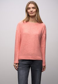 Korallenpinkfarbener Pullover mit lockerem Schnitt, langen Ärmeln und dekorativen Knöpfen an den Schultern. Weiche Textur, kombiniert mit dunklen Jeans.