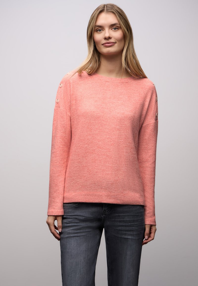 Korallenpinkfarbener Pullover mit lockerem Schnitt, langen Ärmeln und dekorativen Knöpfen an den Schultern. Weiche Textur, kombiniert mit dunklen Jeans.