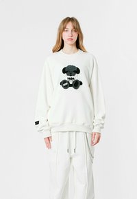 Sudadera blanca con bordado de un oso de peluche negro y gris en el centro. Presenta mangas largas y puños acanalados en el dobladillo. Ajuste casual.