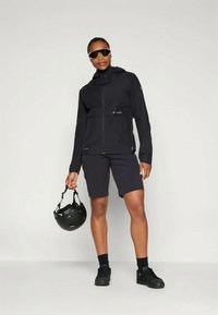 Άτομο ντυμένο με μαύρη ζακέτα ποδηλασίας, σορτς, γυαλιά ηλίου και παπούτσια, κρατώντας ένα μαύρο κράνος ποδηλασίας σε απλή φόντο.