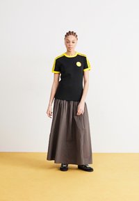 Black t-shirt with yellow accents and a smiley patch, paired with a dark brown skirt, worn with black shoes. Standing on a yellow surface.