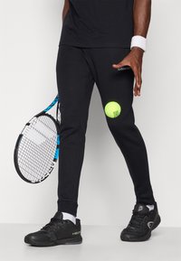 Pantalon de sport noir avec une texture lisse, présentant une balle de tennis posée sur la cuisse, accompagné de chaussures noires et d'une raquette de tennis.