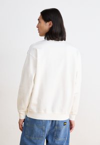 Sweatshirt blanc à coupe décontractée, col et poignets côtelés. Porté avec un jean en denim bleu avec des coutures visibles et une petite étiquette de marque.
