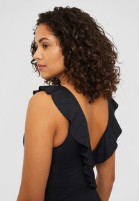 Maillot de bain une pièce noir avec des bretelles à volants, présentant un design à dos en V plongeant et un tissu texturé. Silhouette lisse et ajustée.