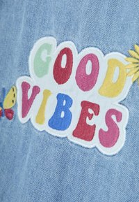 Una tela de denim con el texto bordado colorido "GOOD VIBES." Presenta un diseño de mariposa y flores en tonos brillantes sobre un fondo azul claro.