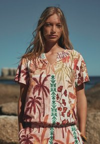 Pitkähiuksinen nainen, joka on pukeutunut trooppiseen kukkamekkoon, seisoo ulkona kivien ja veden edessä kirkkaan taivaan alla.