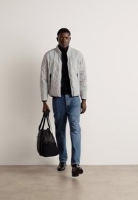 Quilted light gray bomber jacket over a black turtleneck, blue jeans, and black shoes, holding a black bag. Simple design with a textured surface.