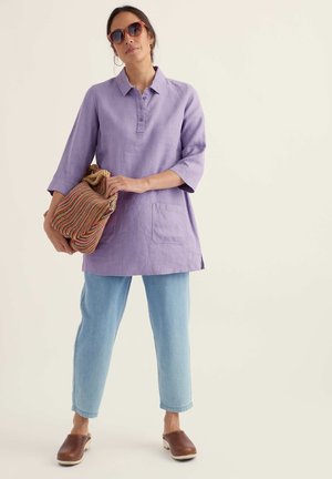 Frau trägt lila Tunika, hellblaue Jeans, braune Clogs, Sonnenbrille und hält eine geflochtene, mehrfarbige Tasche vor neutralem Hintergrund.