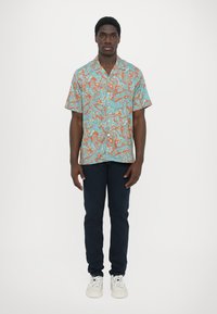 Korte mouwen shirt in turquoise met oranje patronen, met knoopsluiting; gecombineerd met donkerblauwe broeken en witte sneakers.