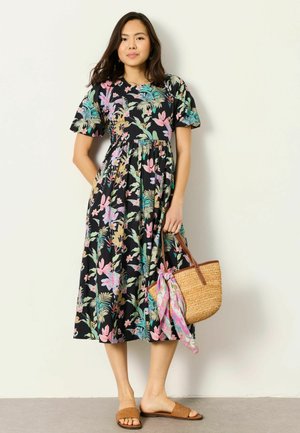 Kvinde iført sort midi-kjole med farverigt blomsterprint, holder flettet halmtaske med pink tørklæde, stående mod en lys væg.