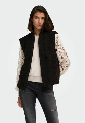 Kvinde iført en sort blød vest over en hvid blondeærmet top og revnede mørkegrå jeans mod en enkel lys baggrund.