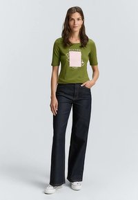 Olivgrünes T-Shirt mit kurzen Ärmeln, einem pinkfarbenen Grafikelement und weißem Text, kombiniert mit dunkelblauen Schlagjeans und weißen Sneakern.