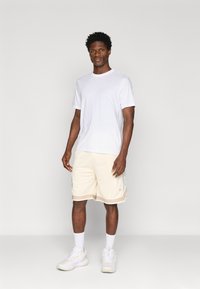 Camiseta branca de manga curta combinada com calções de malha bege com detalhes laterais estampados, usada com ténis brancos e meias até ao tornozelo.