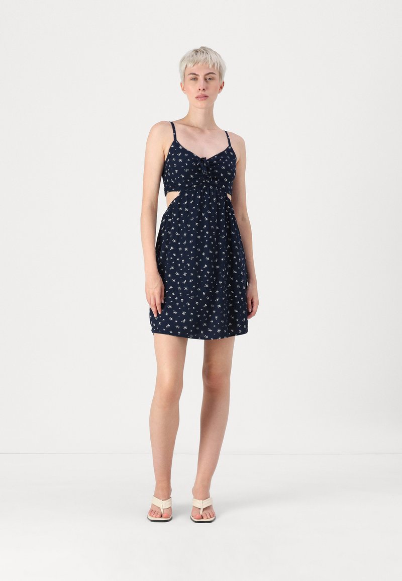 Vestido azul-marinho com pequeno padrão floral branco, corpete ajustado, laterais recortadas e saia rodada. Alças finas, combinado com sandálias leves.