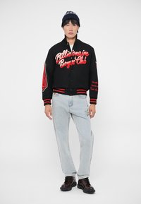 Casaco varsity preto com texto bordado em vermelho, camisa branca, jeans azul claro e sapatos escuros. Gorro com um padrão na aba.