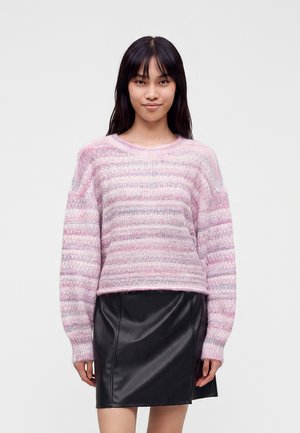 Jeune femme portant un pull en maille rayé rose et gris et une jupe en cuir noire, debout devant un fond clair uni.