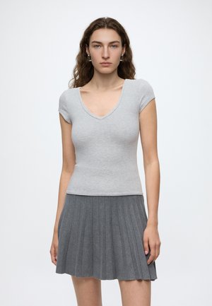 Jeune femme aux longs cheveux bruns bouclés portant un t-shirt ajusté gris clair à col en V et une jupe plissée gris foncé, debout devant un fond uni.