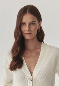 Weißer gestrickter Cardigan mit tiefem V-Ausschnitt und einem einzelnen Holzknopf. Verfügt über ein geripptes Muster und lange Ärmel.