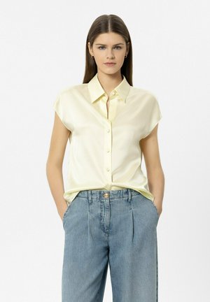 Junge Frau mit glattem braunem Haar trägt ein blassgelbes kurzärmeliges Hemd mit Knöpfen und hoch taillierte lockere blaue Jeans, die Hände in den Taschen.